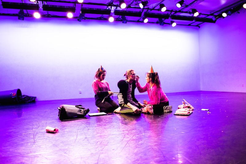 3 dancers sit on the floor wearing party hats. One dancer applies eye makeup to another. A garbage can lays on the ground